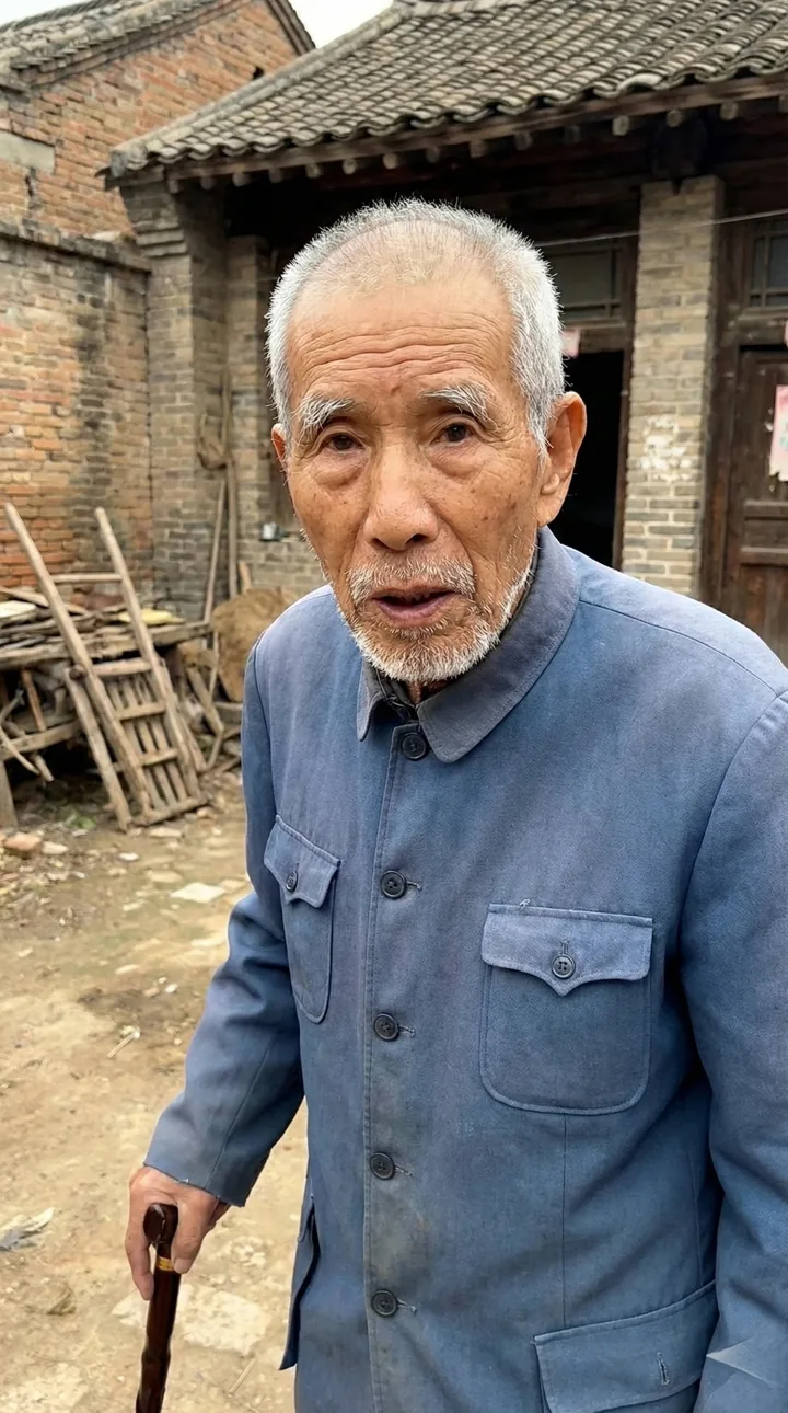 身穿蓝色中山装的老年男性站在乡村老屋前，手持拐杖
