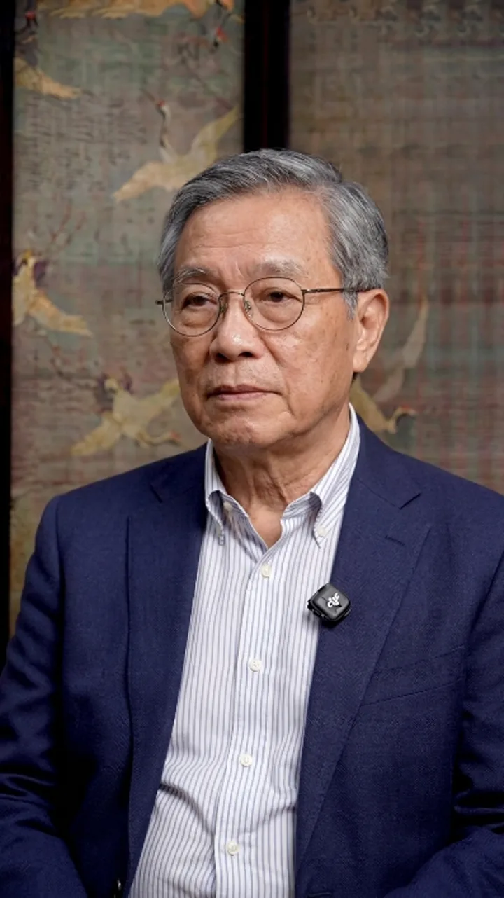 戴圆框眼镜的老年男性穿深蓝西装坐在访谈背景前，佩戴领夹麦克风