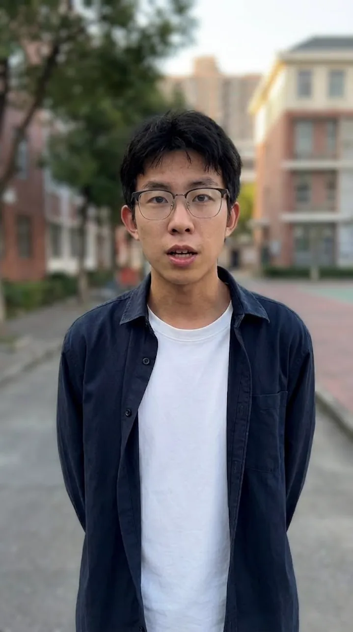 戴眼镜的青年男性站在校园道路上，穿白色上衣和深色外套